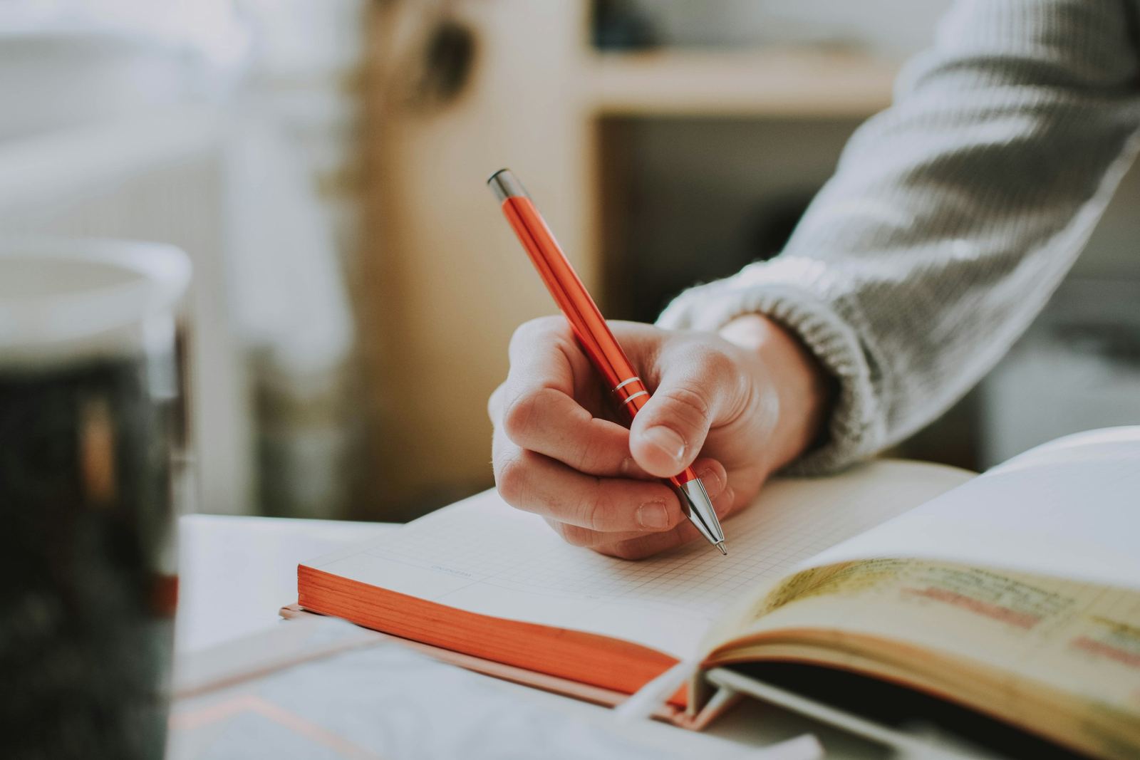Mano escribiendo notas en un cuaderno con una lapicera naranja.