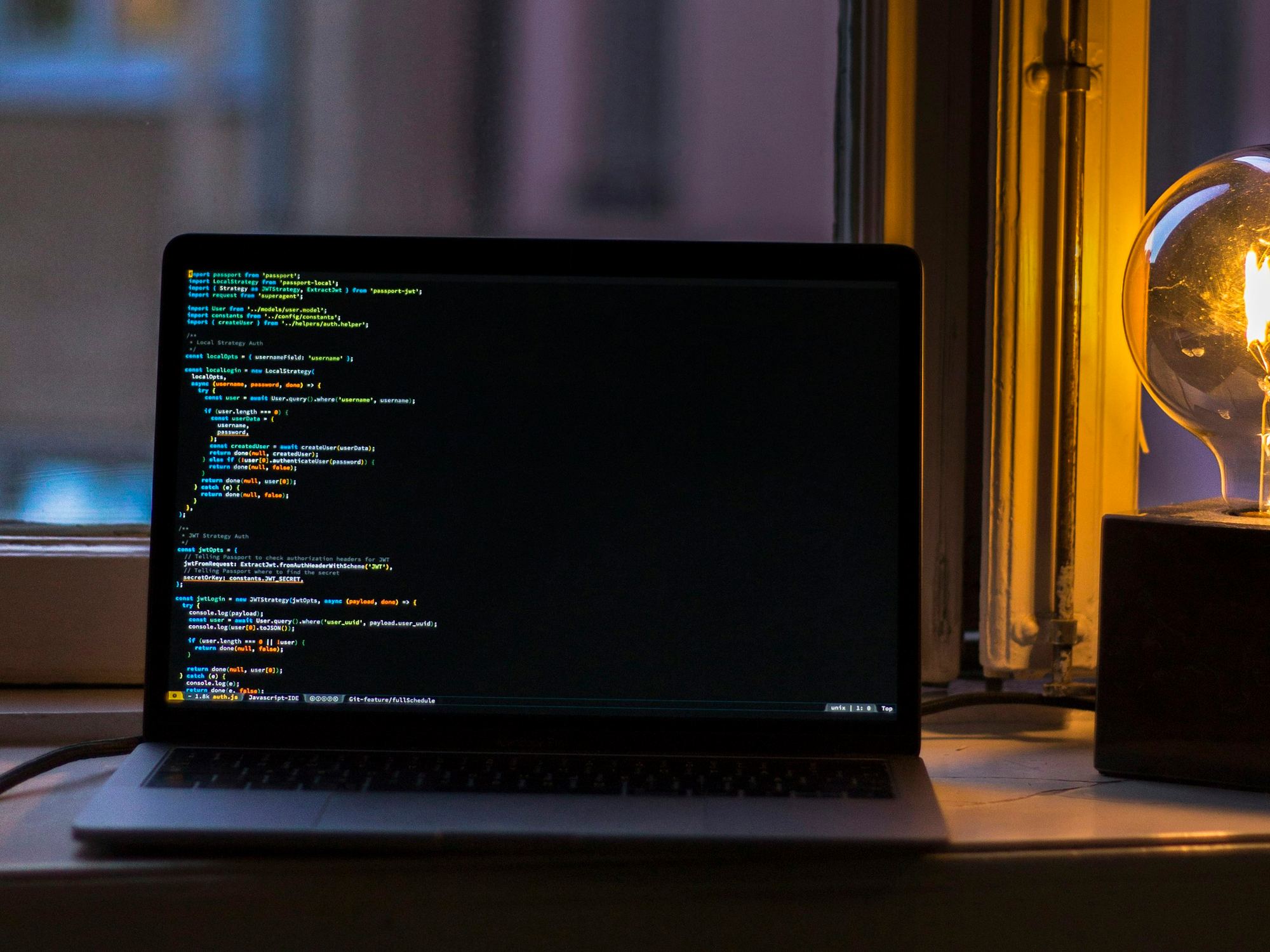 Laptop con código en pantalla junto a una ventana y una lámpara de filamento encendida.